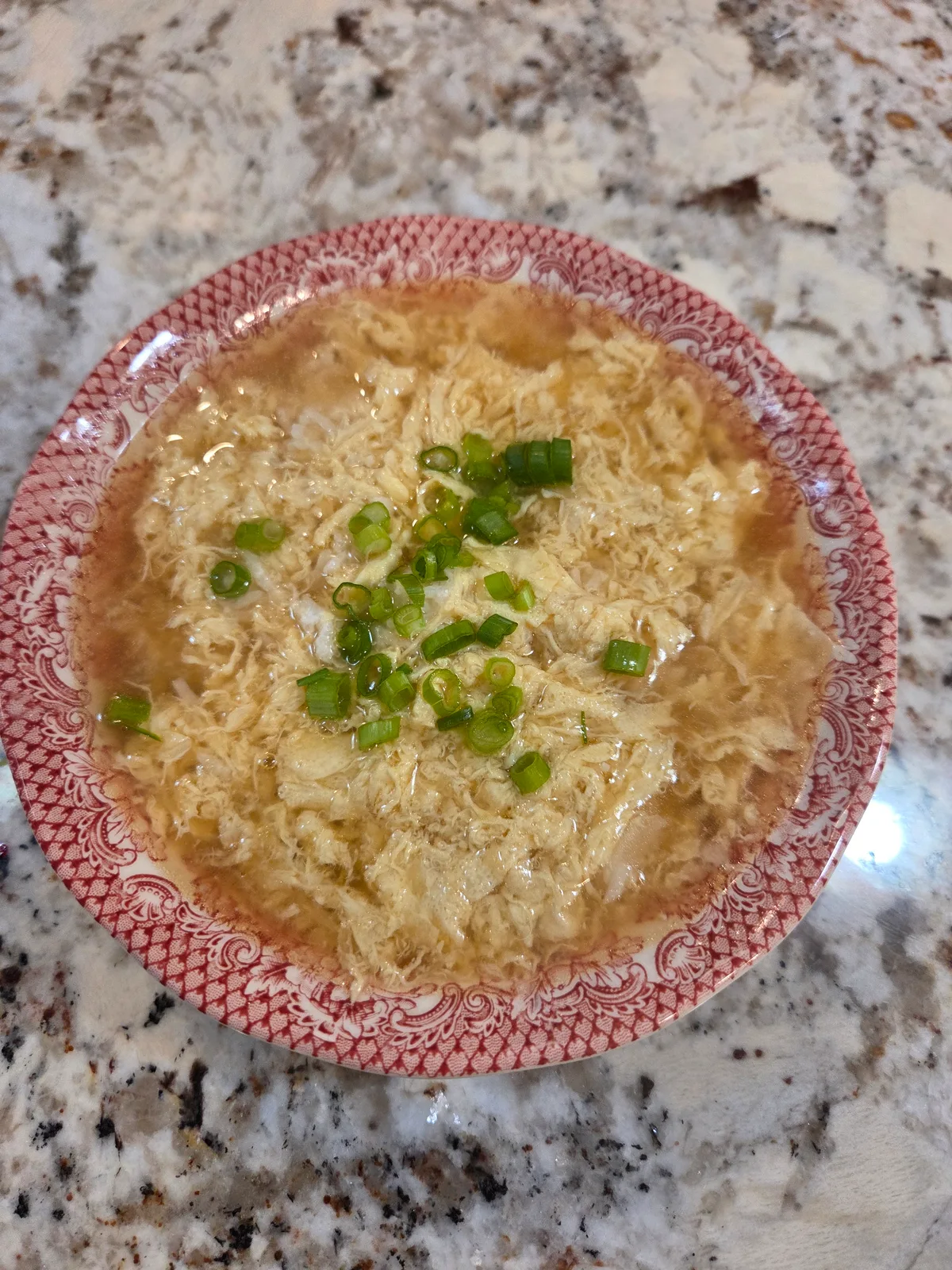 Egg drop soup with egg ribbons and sliced green onions in a red-patterned bowl on a countertop.